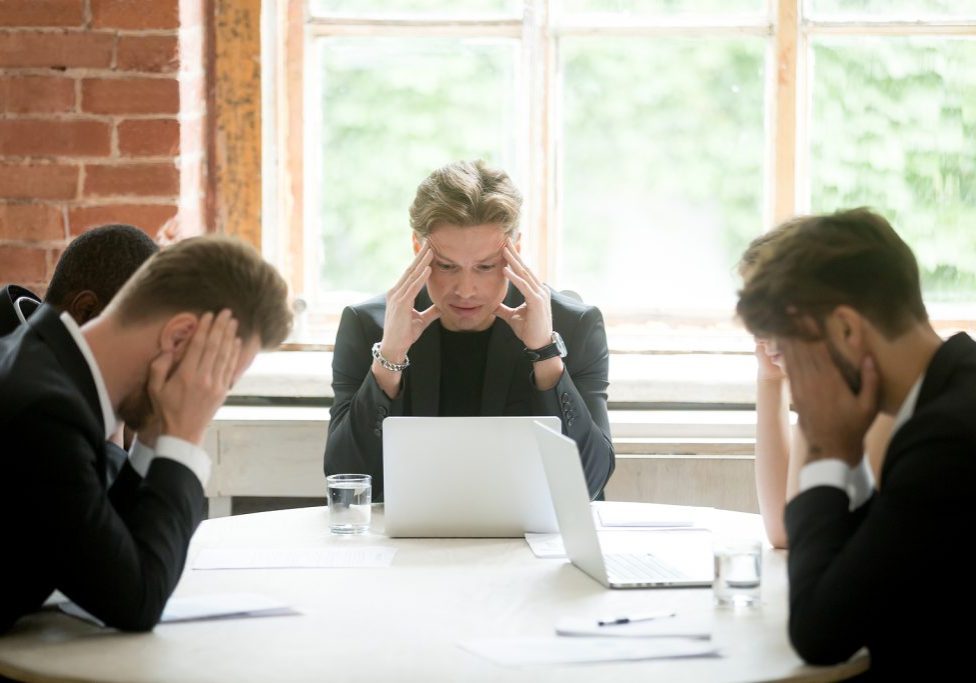 Stressed boss and executive team searching problem solution at meeting, partners holding heads in hands depressed by failure bad news, feeling desperate about company bankruptcy or financial crisis